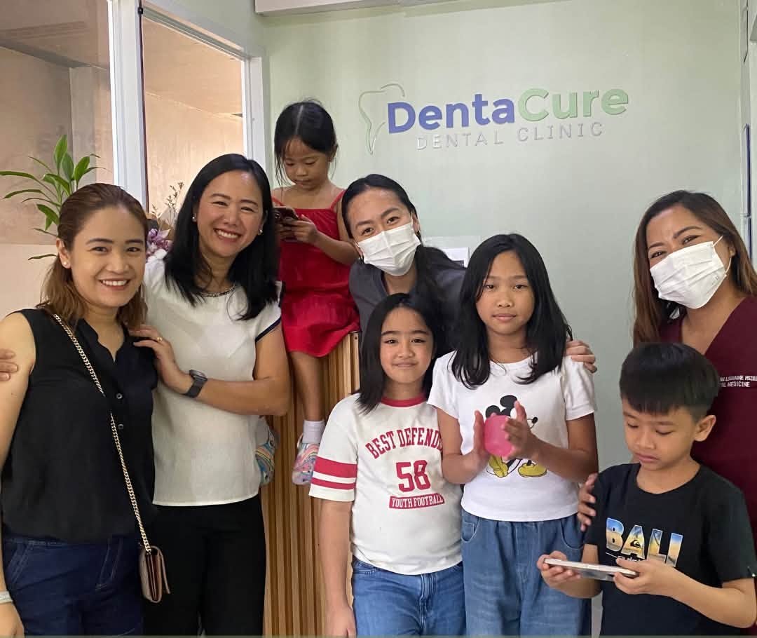 Denta Cure staff and patients smiling together in front of the clinic's logo wall